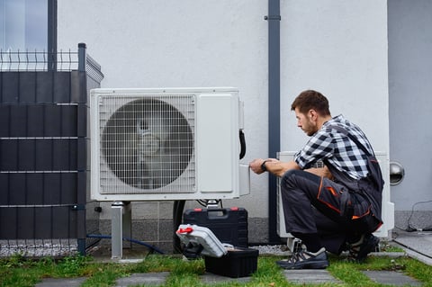 Technician performing maintenance on outdoor heat pump unit near residential building exterior